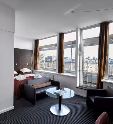 Hotelværelse med stort vinduesudsigt over byens bygninger. Værelset har en dobbeltseng med hvidt og rødt sengetøj, et rundt glasbord, en sofa og stole. Døren har teksten "Square Lounge" trykt på. Værelset er veloplyst og moderne.