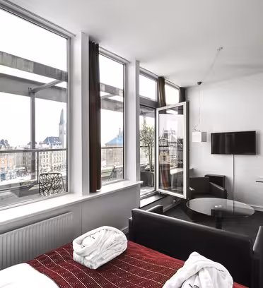Et moderne hotelværelse med et stort vindue med udsigt over bybilledet. Værelset har en seng med en rød mønstret løber, en sofa, to badekåber og et væghængt tv. Indretningen er minimalistisk med to sort-hvide billeder på væggen.