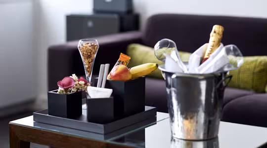 Et hotelværelsesopsætning med et glasbord med et frugtfad, chokolade og jordnødder i sorte beholdere. En champagneflaske køler i en isspand. I baggrunden ses en mørk sofa med grønne hynder.