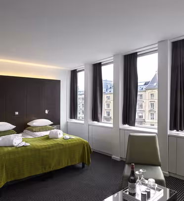 Moderne hotelværelse med en dobbeltseng dækket af et grønt tæppe, puder og håndklæder. Store vinduer med mørke gardiner giver udsigt over byen. Et glasbord rummer vandflasker og glas. Hvide vægge og mørkt tæppe tilføjer et slankt touch.