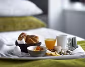 En morgenmadsbakke på en seng med en kurv med croissanter, en kop kaffe, et glas appelsinjuice, et kogt æg, en skål frugt, kød i skiver, ost og agurk. Det grønne mønstrede sengetøj ses i baggrunden.