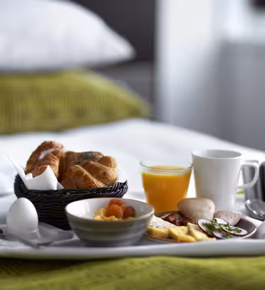 En morgenmadsbakke på en seng med en kurv med croissanter, en kop kaffe, et glas appelsinjuice, et kogt æg, en skål frugt, kød i skiver, ost og agurk. Det grønne mønstrede sengetøj ses i baggrunden.