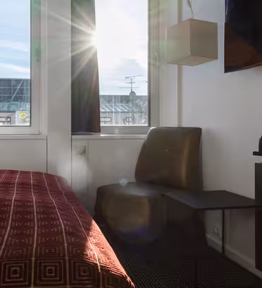 Sollys strømmer gennem et vindue på et moderne hotelværelse og oplyser et rødbrun sengetæppe med geometriske mønstre. En stol og et lille bord er ved siden af et vindue. På den modsatte væg er der monteret tv og kaffestation.