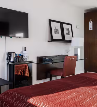 Hotelværelse med en pænt redt seng med et rødt tæppe, et vægmonteret fladskærms-tv og et skrivebord med en rød stol. Sort-hvide indrammede billeder er over skrivebordet ved siden af en lampe og gratis drinks på en bakke. Et skab er i hjørnet.
