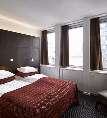 Moderne hotelværelse med dobbeltseng, mørkt polstret sengegavl og rødmønstret sengetæppe. To store vinduer med brune gardiner giver udsigt til en bybygning. En brun stol sidder til siden på et tæppebelagt gulv.