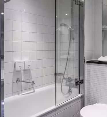 Moderne badeværelse med hvide flisevægge med badekar med bruseniche af glas. Der er to sæbedispensere på væggen ved siden af karret. Et toilet og rullede hvide håndklæder på en hylde er synlige til højre.