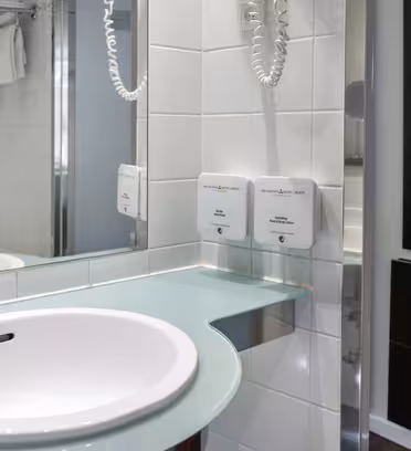 En moderne badeværelsesvask med en cirkulær hvid vask sat ind i en glasbordplade. I nærheden er en monteret hårtørrer og sæbedispensere. Rummet har hvide flisevægge og et spejl med refleks. Håndklæder er stablet på en hylde ovenover.