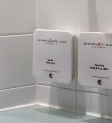 Vægmonterede dispensere i badeværelse, mærket "Gentle Hand Wash" og "Hydrating Hand & Body Lotion" fra Arp-Hansen Hotel Group, København. De er placeret over en grøn bordplade med flisebelagte vægge.