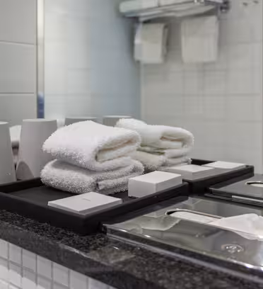 Hotelbadeværelsesdisk med pænt foldede hvide håndklæder, hvide kopper, sæbe og papirservietter på en mørk bordplade. Et stort spejl reflekterer en del af rummet, og en matteret glasvæg med synlig tekst er i baggrunden.