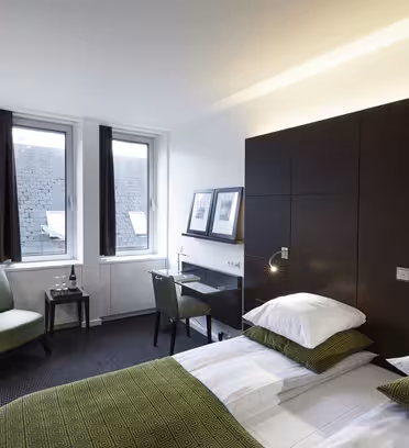 Et moderne hotelværelse med en pæn opredt seng med grønne puder og et grønt læg. Der er et lille skrivebord med en stol, en grøn lænestol, to indrammede billeder og to vinduer, der giver naturligt lys ind i rummet.