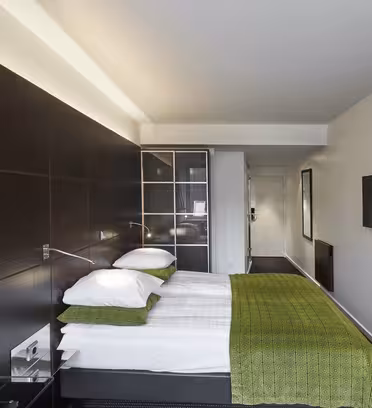 Moderne hotelværelse med en pænt lavet dobbeltseng med grønne accenter. Rummet har hvide vægge, et væghængt tv, en stor sengegavl i mørkt træ, indrammet kunst på en hylde og et lukket skab. Rummet er lyst og minimalistisk.