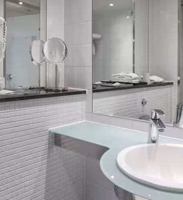 Moderne badeværelse med hvide flisevægge og stort spejl. Har en hvid vask med et elegant vandhane, glasbordplade, hårtørrer, foldede håndklæder og toiletartikler. Toiletpapirholder og en håndklædeholder med foldede hvide håndklæder er synlige.