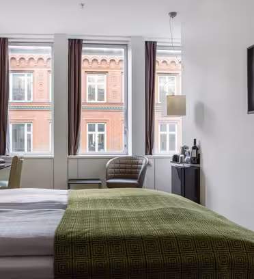 Et moderne hotelværelse med en pæn opredt seng med grønt sengetøj. Et fladskærms-tv er monteret på væggen. Store vinduer afslører udsigten til en rød murstensbygning udenfor. Værelset indeholder en stol og et skrivebord med en lampe.