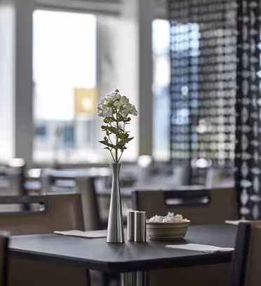 Et moderne, tomt restaurantinteriør med fokus på et bord. Bordet er dækket med en metalvase med hvide blomster, en skål og krydderier. Blød belysning filtrerer gennem store vinduer, og geometriske mønstre dekorerer en skillevæg i baggrunden.