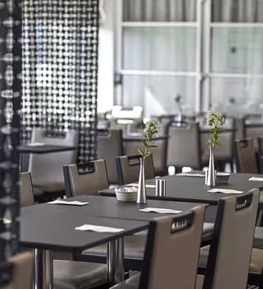 Et moderne restaurantinteriør med pænt arrangerede borde med sorte overflader. Hvert bord har en enkel metalvase med små blomster. Rummet er opdelt af sorte geometrisk-mønstrede skærme, og store vinduespartier giver naturligt lys.