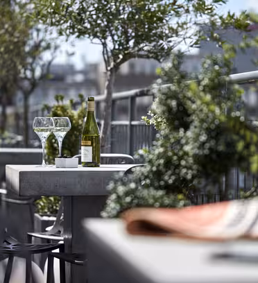 En tagterrasse med et betonbord med to tomme vinglas og en flaske vin. Frodigt grønt omgiver området, og et bybillede er sløret i baggrunden. En foldet avis og kaffekop sidder ude af fokus i forgrunden.