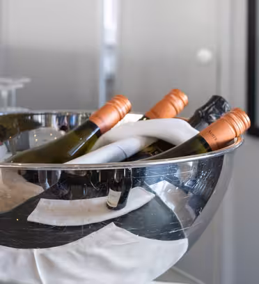 En sølvskål fyldt med flere vinflasker, hver med orange eller sorte toppe, er foret med en hvid serviet. En flaske står uden for skålen til højre, og flere tomme glas er sløret i baggrunden.