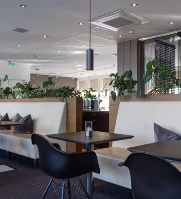 Moderne caféinteriør med polstrede kabinesæder, sorte borde og slanke sorte stole. Grønne planter pryder skillevæggene. Neutrale toner dominerer indretningen og skaber en minimalistisk og hyggelig atmosfære.
