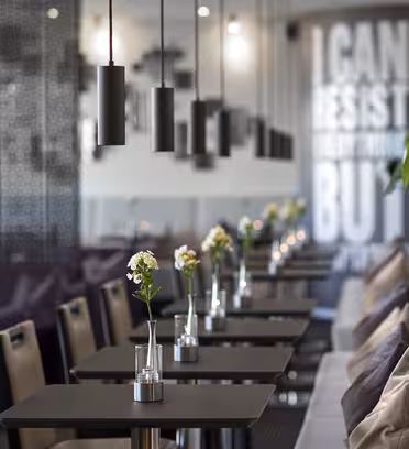 Et moderne cafeinteriør med en række firkantede borde og stole. Hvert bord har en lille vase med en blomst. Sorte pendellamper hænger fra loftet. Der er en væg med teksten "I CAN RESIST EVERYTHING BUT..." og en række planter over en polstret bænk.