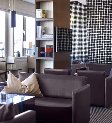 Moderne loungeområde med mørkebrune sofaer og borde med glasplade. En central skillevæg med hylder viser bøger og indretning. Værelset har stilfulde sorte og hvide geometriske mønstre på væggene, med store vinduer, der slipper naturligt lys ind.