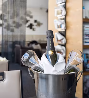 En flaske champagne i en metalisspand er omgivet af fire tomme glas på et bord i en eksklusiv restaurant. I baggrunden er der en moderne indretning med siddepladser, bogreoler og abstrakt vægkunst.