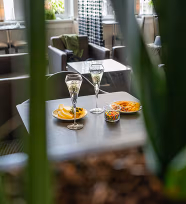 To glas champagne på et bord med små tallerkener chips og en skål med farverige snacks. Scenen ses gennem grønne planteblade med hyggelige café siddepladser i baggrunden.