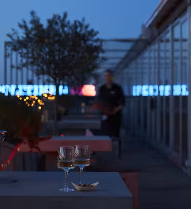 Rooftop i skumringen med to glas hvidvin på et bord. En flaske i en isspand og et lille fad ledsager dem. Der er varm strengbelysning, og en sløret figur er i baggrunden. Skilte er synlige i det fjerne.