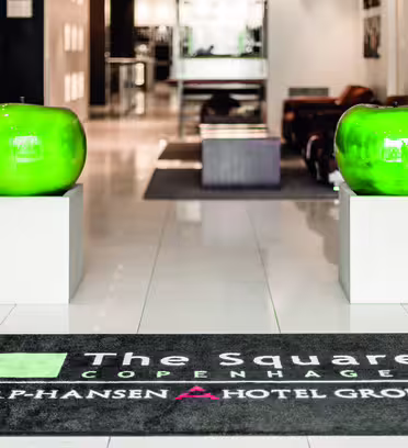 Indgang til The Square Hotel i København, med to store grønne skulpturer på piedestaler og en mærkevaregulvmåtte. Interiøret har et slankt, moderne design med et lyst, poleret gulv og blød belysning.